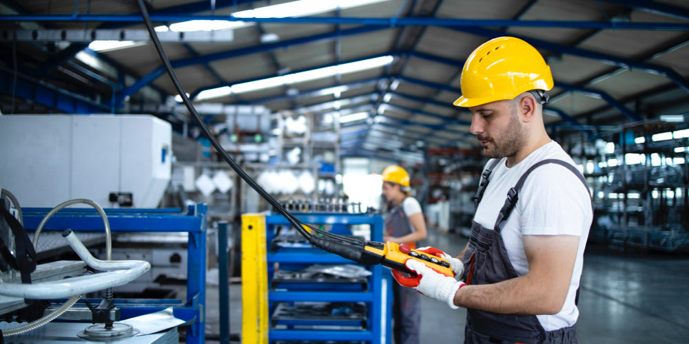 factory worker wearing uniform hardhat operating industrial machine with push button joystick production hall 1000x500